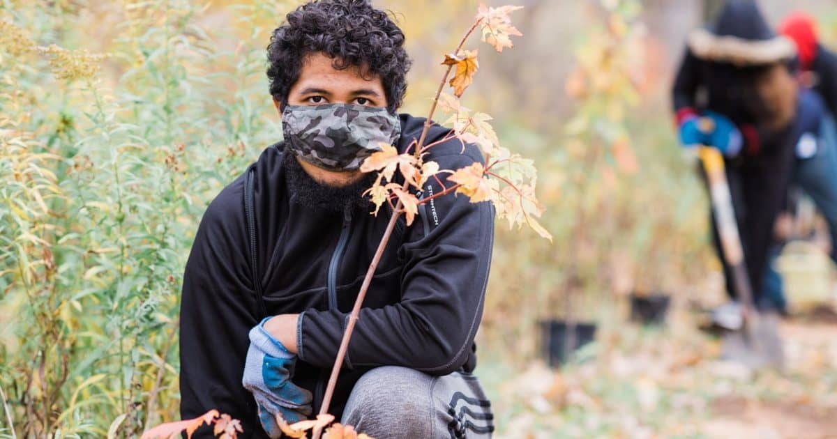 a student wearing work gloves and a mask kneeling behind a sapling they planted with other students working in the background