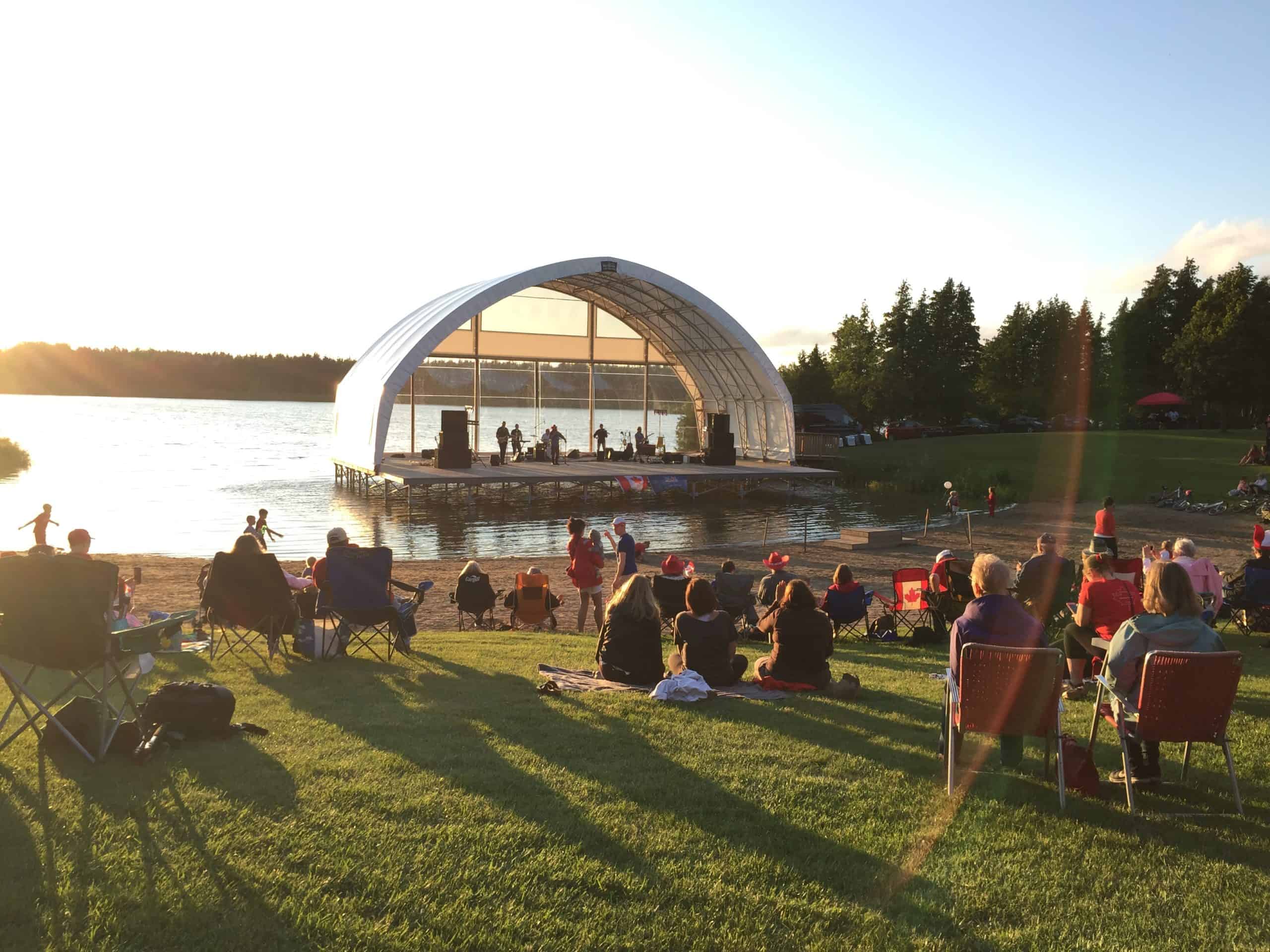 People sitting in front of an ampthitheatre with a band playing on the stage and water in the background