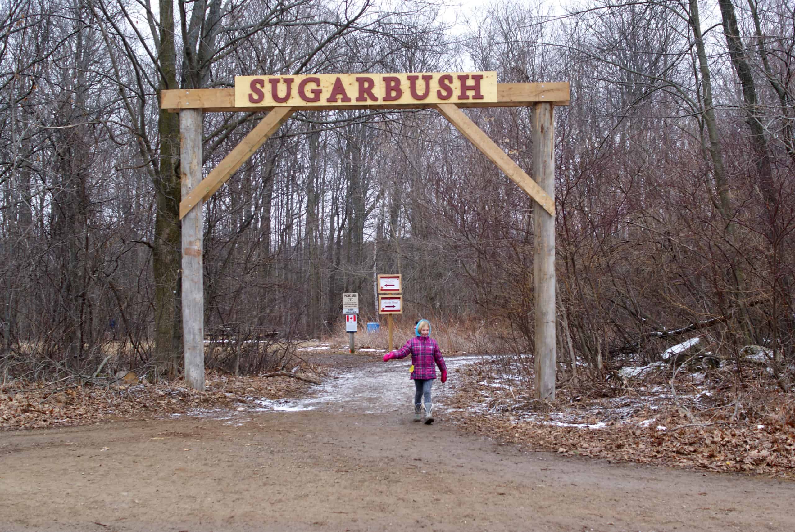 archway with Sugarbush on it and a young person walking through it with trees in the background